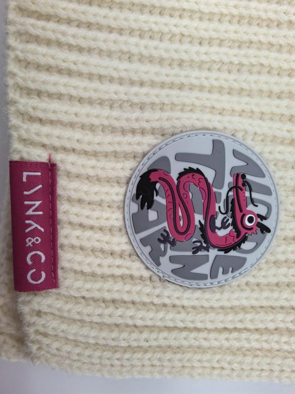 Cream chunky ribbed knit beanie with pink dragon circular rubber patch and pink brand label on textured wool fabric
