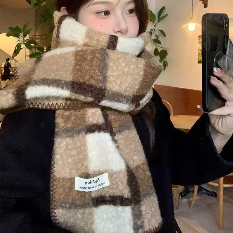 Young woman wearing a thick plaid fuzzy scarf in beige, brown and cream tones, taking a selfie indoors