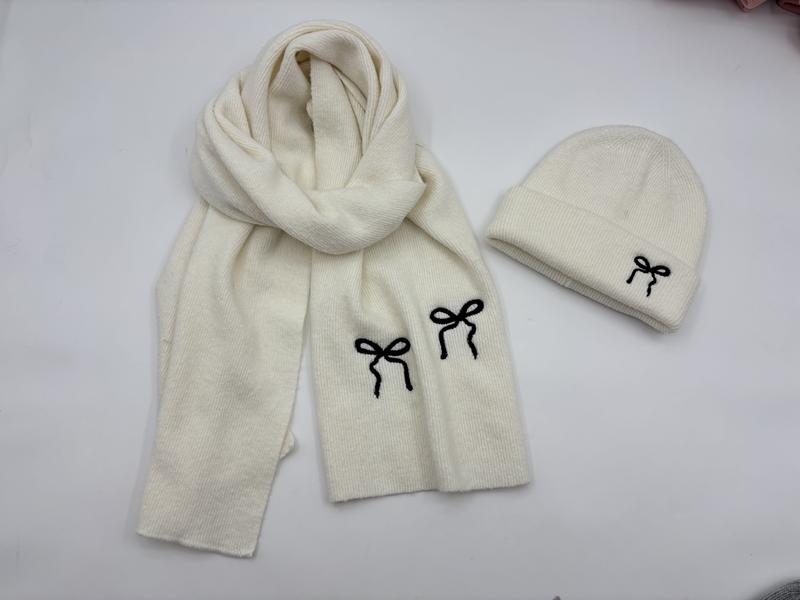 Cream ribbed knit winter hat and scarf set with black embroidered bow details laid flat on white surface