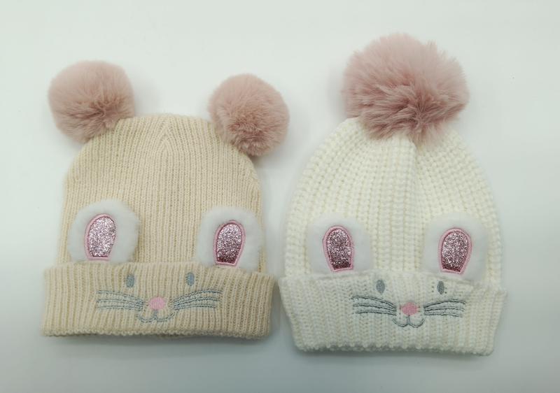 Two knit baby beanies in cream and white with glitter bunny ear patches, embroidered whiskers, and pink faux fur pompoms on top
