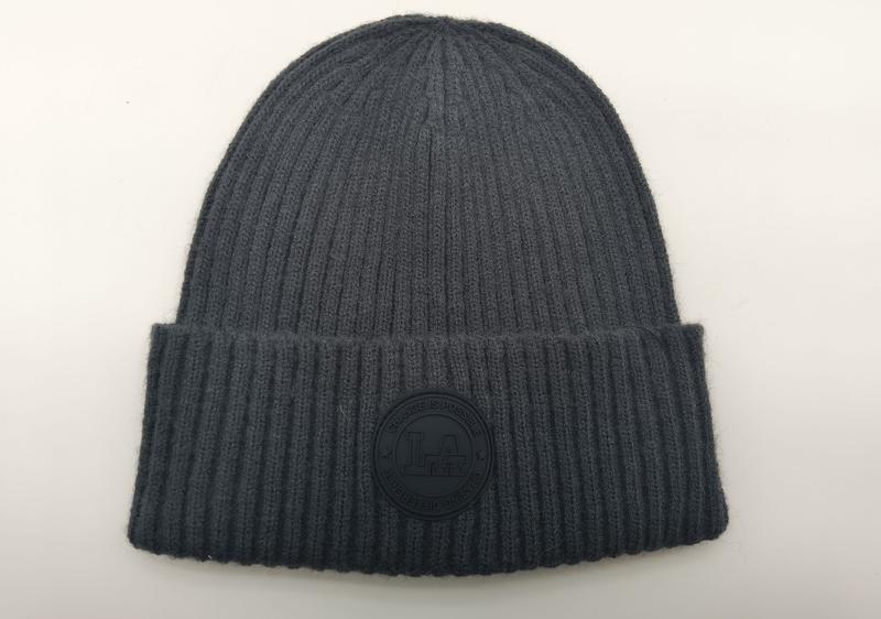 Charcoal grey ribbed knit cuffed beanie hat with circular rubber logo patch reading Change Is Possible, laid flat on white background