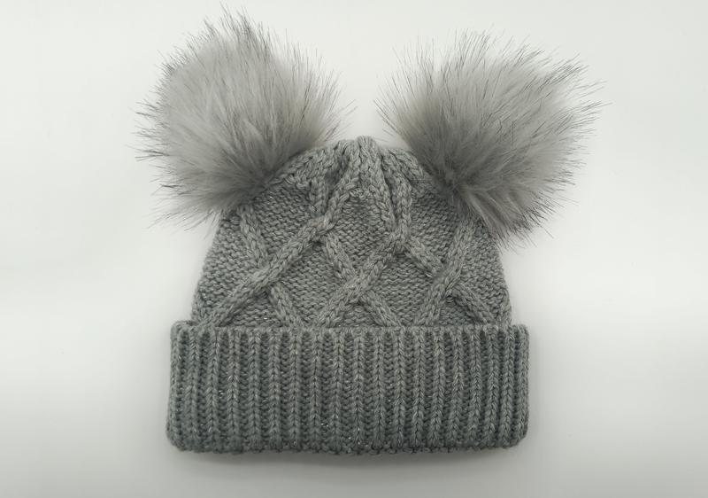 Gray cable knit winter beanie hat with two fluffy faux fur pom poms on top, ribbed cuff, flat lay on white background