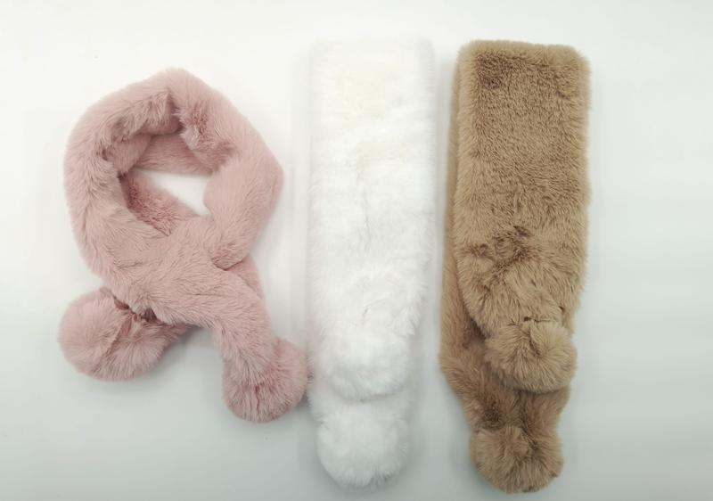 Three fluffy faux fur pull-through scarves with pom pom ends in blush pink, white, and tan laid flat on white background