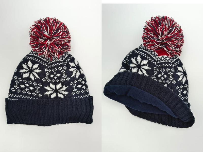 Navy blue knit beanie hat with white Fair Isle snowflake pattern, red and white multicolor pom-pom, and fleece-lined interior shown front and side view