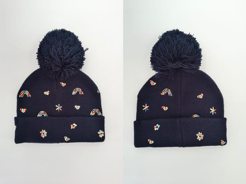 Navy blue knit beanie hat with large pom pom and colorful embroidered rainbows, hearts, stars and flower motifs, shown front and side view