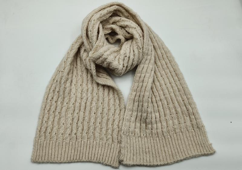Cream beige cable knit scarf loosely coiled on white background showing textured pattern and ribbed hem ends