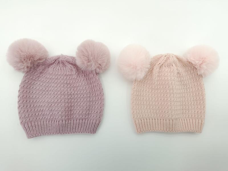 Two knitted winter beanies with double faux fur pom-poms, one in mauve purple and one in blush pink, on white background