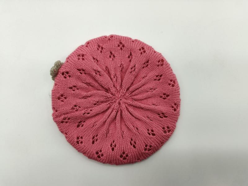 Flat lay of a pink knitted beret with radiating lace floral eyelets and a small tan pompom on a white background