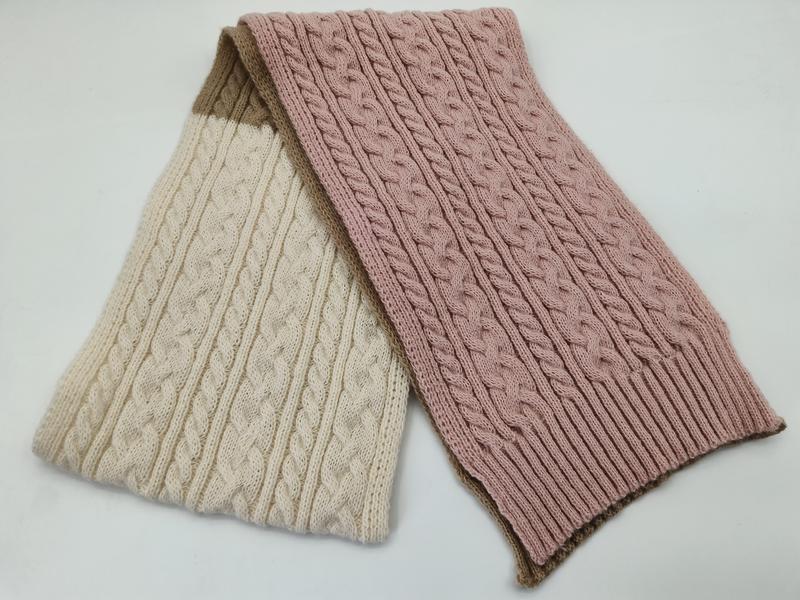 Two cable knit scarves folded together, one cream and one dusty pink, with tan ribbed edges on white background