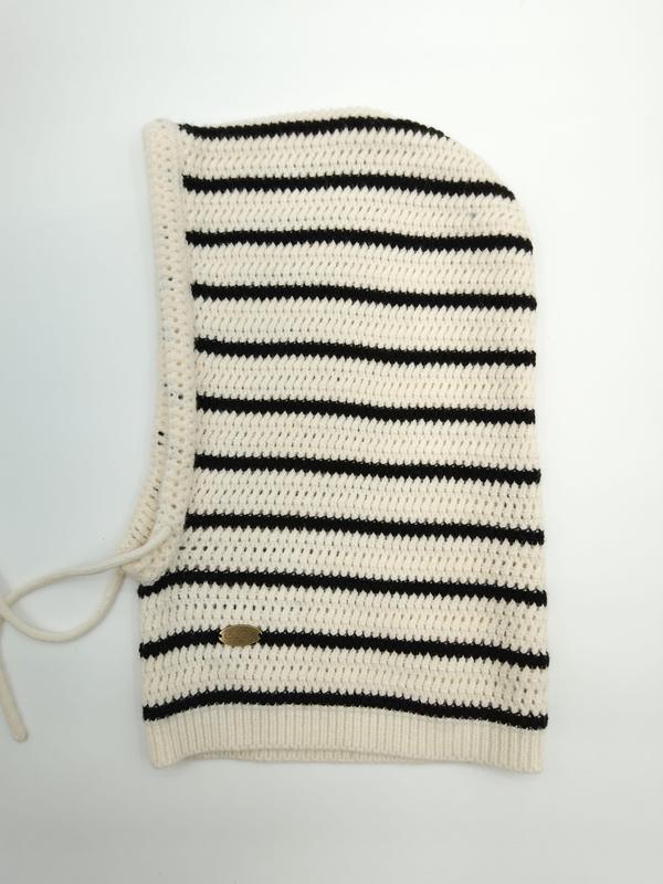 Cream and black striped crochet knit balaclava hood laid flat, showing ribbed hem, tie closure, and small gold brand label