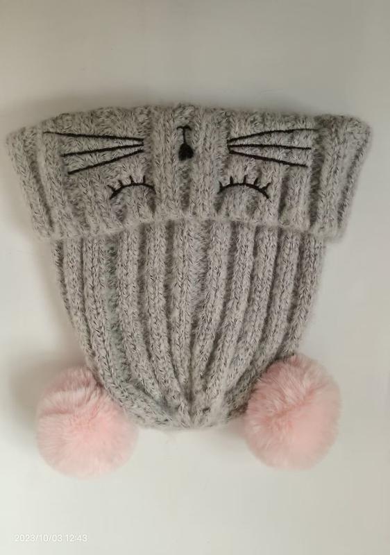 Grey ribbed knit winter beanie hat with embroidered cat face, whiskers, nose, and two pink fluffy pom poms at the base