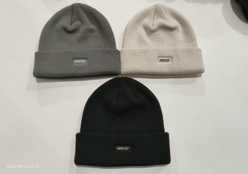 Three ribbed knit cuffed beanies in charcoal grey, cream beige, and black laid flat on white surface with logo patch