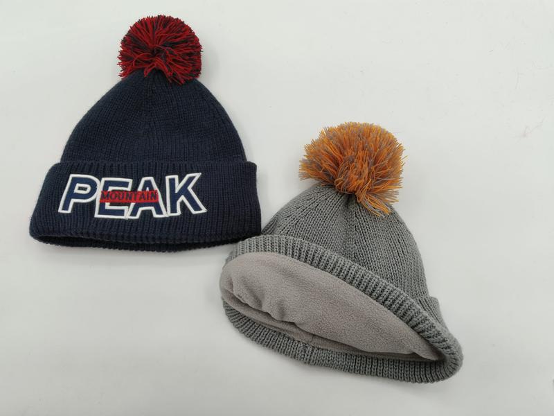Two knit pom-pom beanies: navy blue with Peak Mountain logo and red pom, grey fleece-lined hat with orange pom, on white background