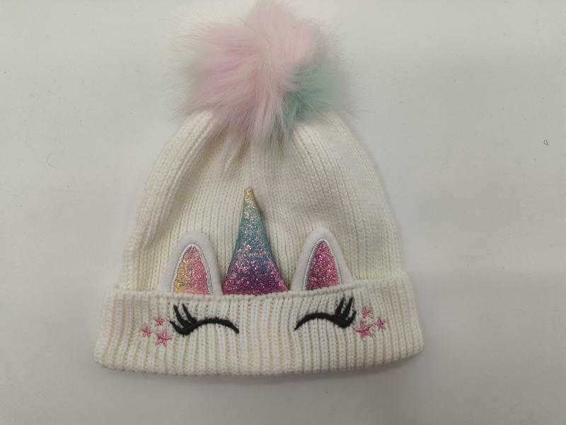 White ribbed knit children's unicorn beanie with glitter horn, pink ears, eyelashes, stars, and pastel rainbow faux fur pom-pom on top