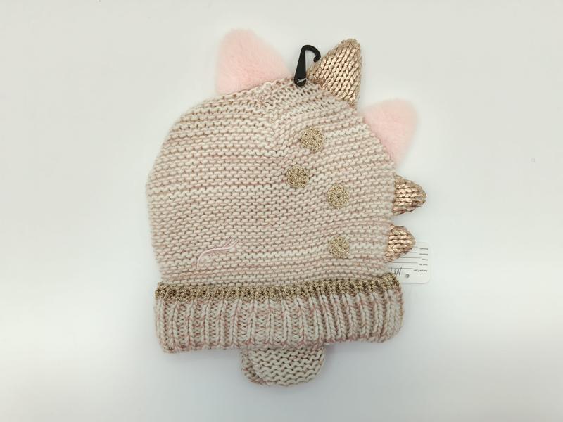 Children's cream knit dinosaur beanie hat with rose gold metallic spikes, pink pompom ears, and gold crochet flower accents on white background