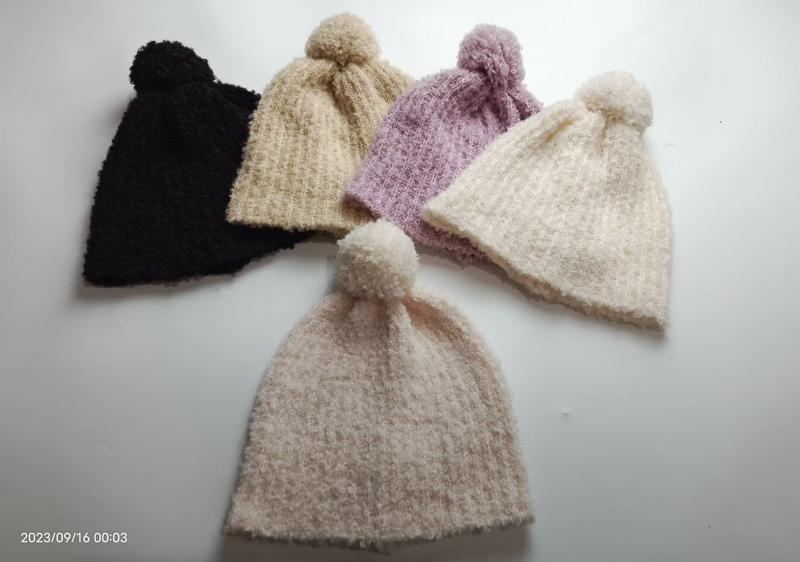 Five soft knitted pom-pom beanies in black, beige, lavender, cream and blush pink arranged on white surface