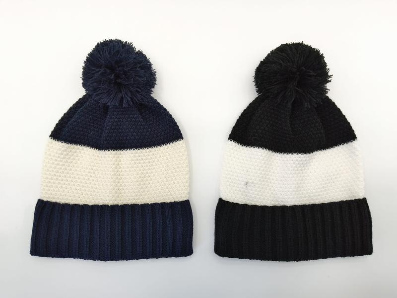 Two knit pom-pom beanies side by side: navy blue and cream stripe on left, black and cream stripe on right, on white background