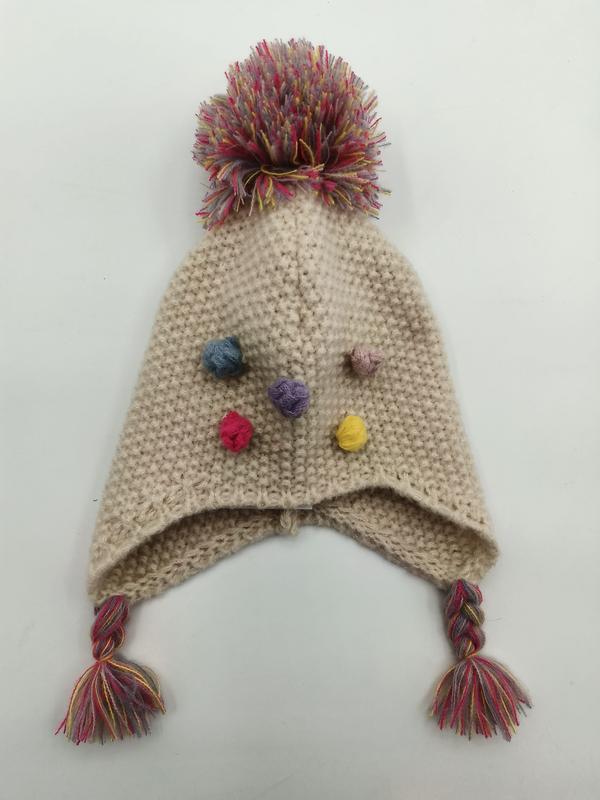 Cream knitted earflap hat with multicolor pompom on top, small colorful yarn bobbles, and braided tassels with fringe ends