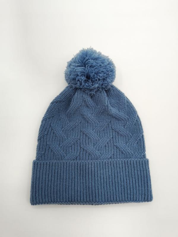 Steel blue cable knit winter beanie hat with fluffy pom pom on top and ribbed cuff, laid flat on white background