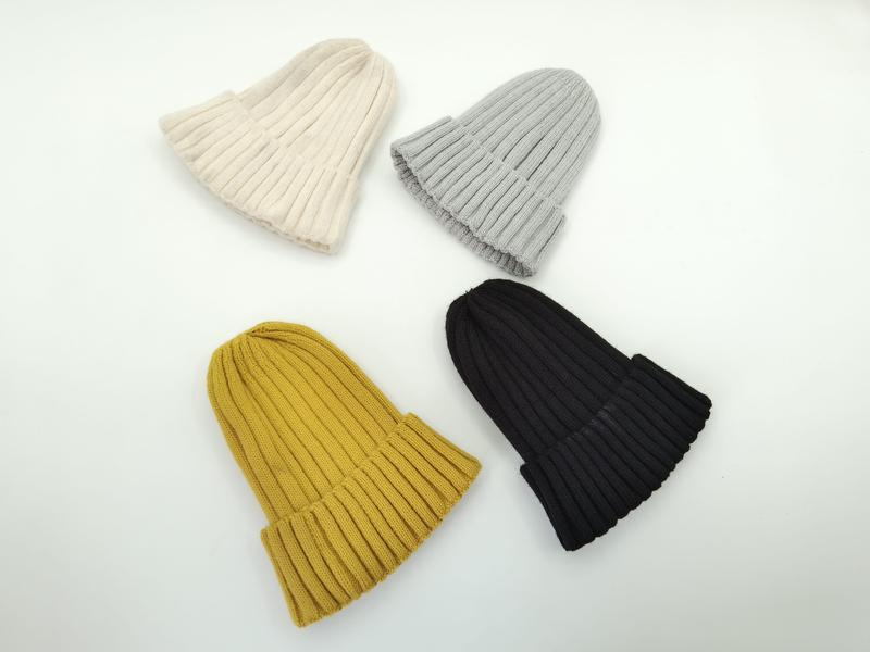 Four ribbed knit beanie hats laid flat in cream, light grey, mustard yellow, and black colorways on white background