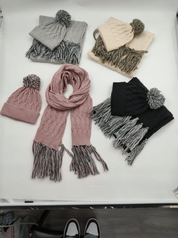Four cable knit winter hat and scarf sets in gray, cream, pink, and black with pom pom beanies and fringed scarves laid flat