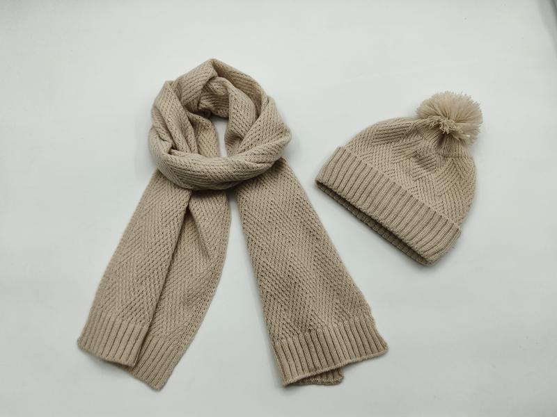 Beige chunky knit scarf coiled and matching pom-pom beanie hat set on white background