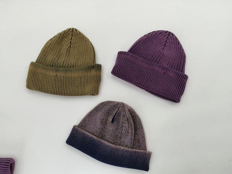Three ribbed knit beanie hats laid flat in olive green, dusty purple, and two-tone gray-navy colorways