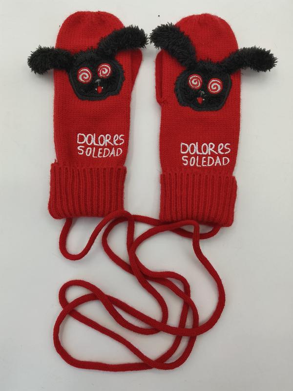 Red knitted mittens with black fuzzy monster face appliqué, spiral eyes, and long connecting cord, laid flat on white background
