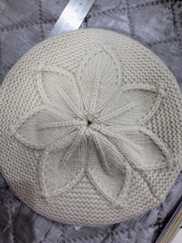Top view of a cream knitted beret with a raised petal flower cable pattern at the crown, laid flat on gray quilted fabric