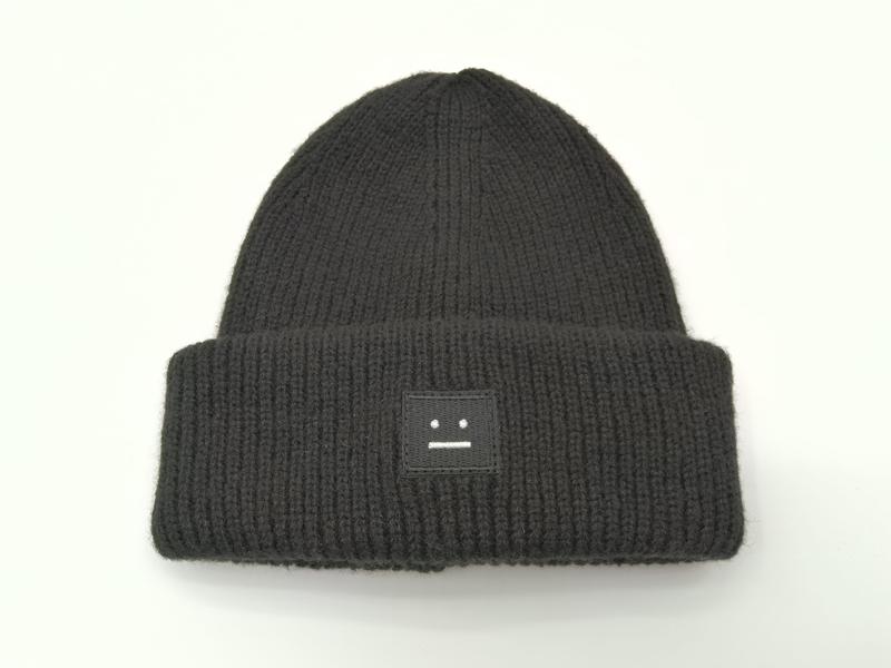 Black chunky ribbed knit beanie hat with folded cuff and small square embroidered neutral face logo patch on white background