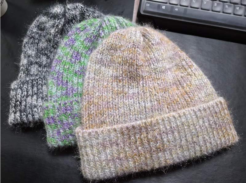 Three handknit wool blend beanie hats in gray marled, green purple stripes, and beige gold tones on dark surface