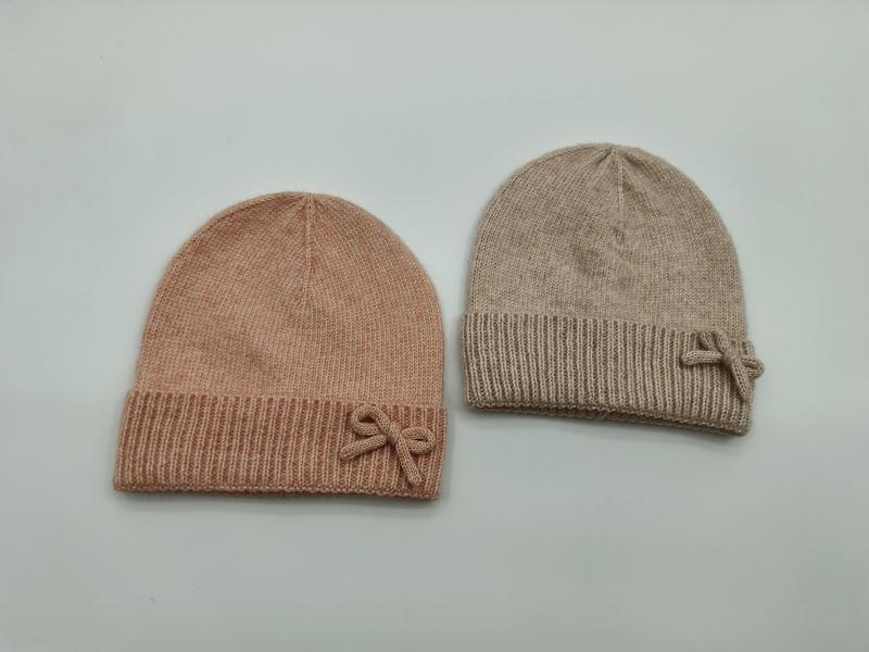 Two soft knit beanie hats with bow accents laid flat, one blush pink and one beige, ribbed cuff design