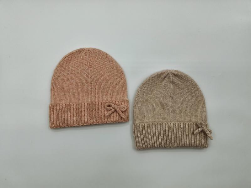 Two soft knit beanie hats with ribbed cuff and bow detail in blush pink and beige, laid flat on white background