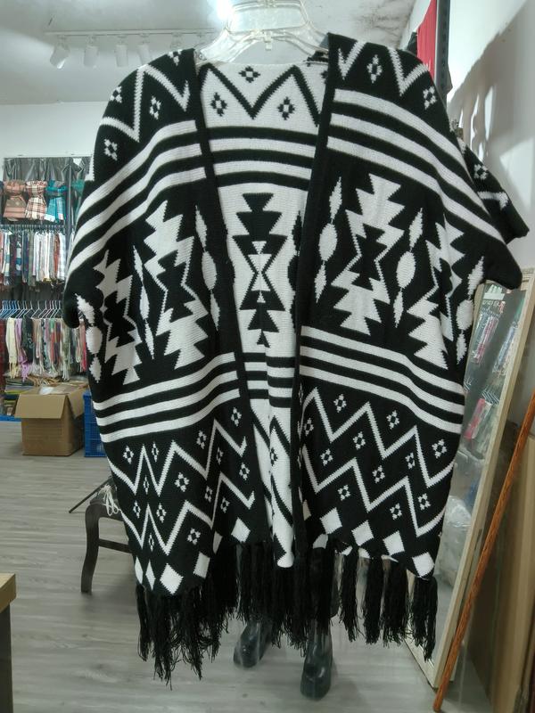 Black and white Aztec geometric pattern knit poncho cape with tassel fringe hem displayed on hanger in store