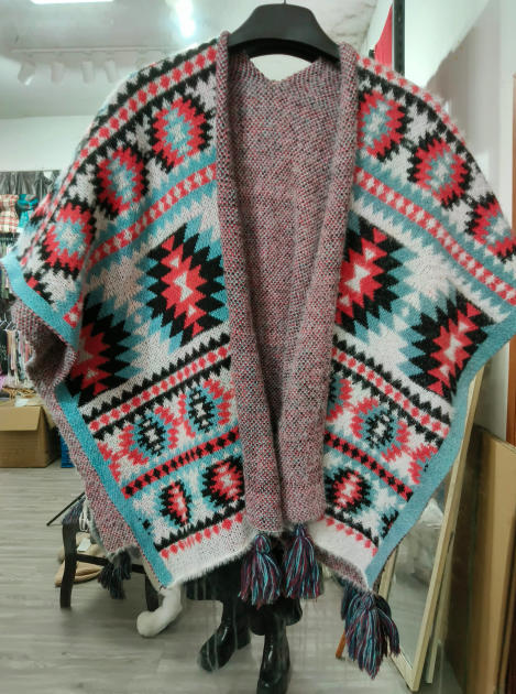 Open-front knitted poncho with Aztec geometric pattern in red, teal, black and white, with multicolor fringe tassels, displayed on hanger