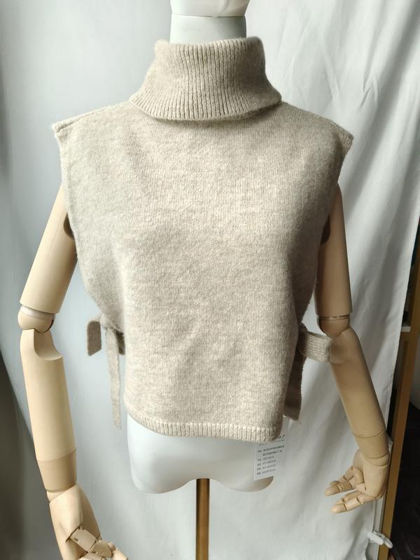 Beige knit sleeveless turtleneck sweater vest with side tie details displayed on a mannequin, soft wool blend
